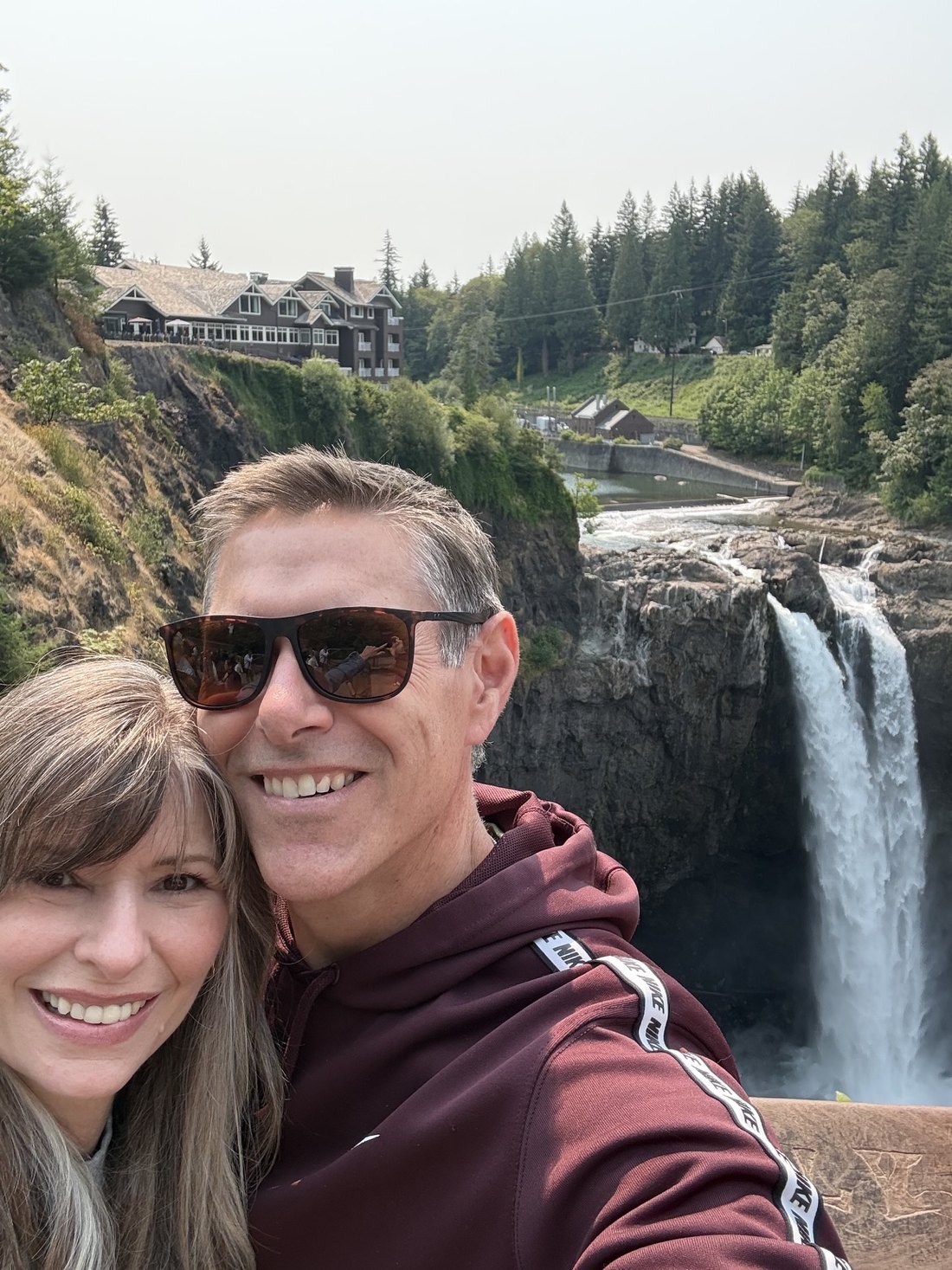 Ann and her husband Al, smiling together outdoors with a waterfall and evergreen trees behind them.